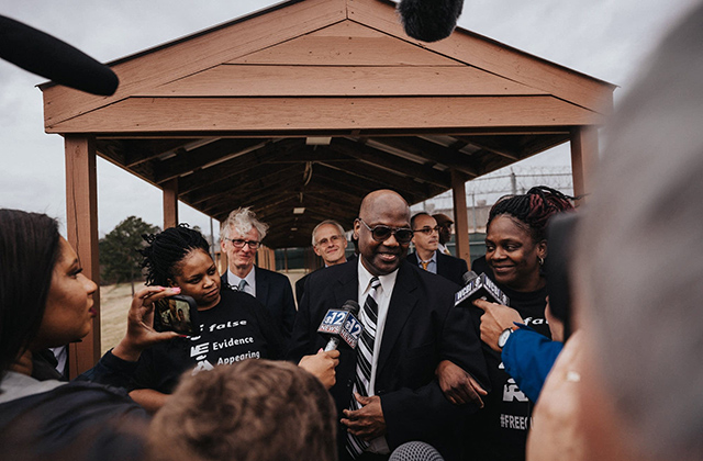 Curtis Flowers. Black man with bald head, dark glasses, dark suit, white shirt and black-and-white tie surrounded by people and reporters.