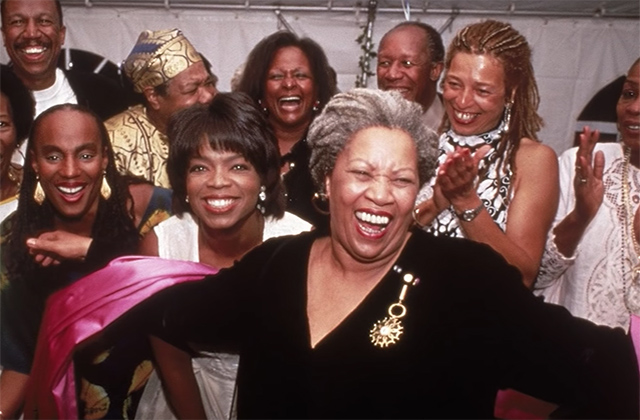 Toni Morrison. Older Black woman with gray locs pulled back wearing black top and surrounded by clapping Black women.