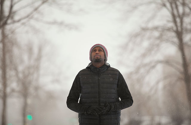 Black man with beard standing in snowy, tree-lined street wearing knit cap, black winter coat, with hands in pockets.