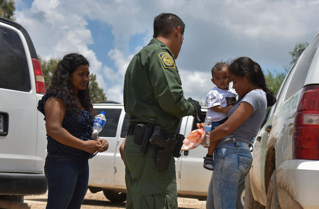 One woman holding an infant and another woman with dark hair stand next to a border patrol agent wearing a green uniform