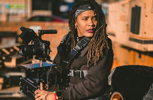 Firelight. Black person with long brown locs wearing black clothing with a black headband, holding  a large camera.
