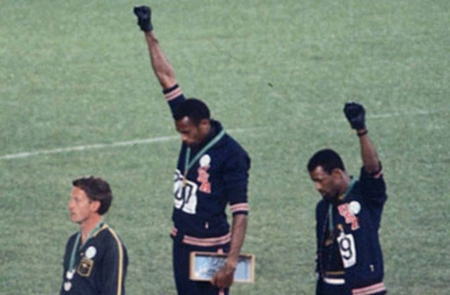 John Carlos and Tommie Smith. Two Black men, at Olympic podium, with black gloved fists raised.