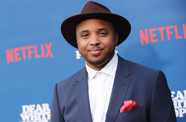 Justin Simien. Black man wearing brown fedora, blue suit jacket and white shirt, standing in front of Netflix signage.