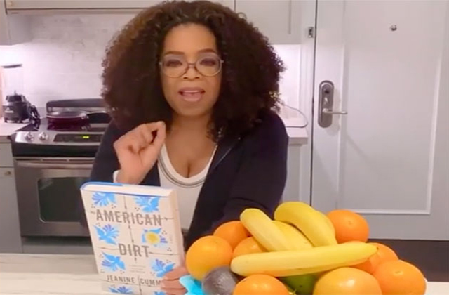 Oprah Winfrey. Black woman with shoulder length curly brown hair wearing cream top and black cardigan, standing at a kitchen counter in front of a fruit bowl, while holding the book "American Dirt."