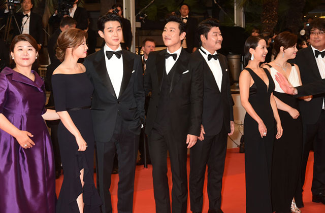 Cast of Parasite. Asian men and women standing on red carpet with one person wearing a purple gown and everyone else dressed in black gowns and tuxedos.
