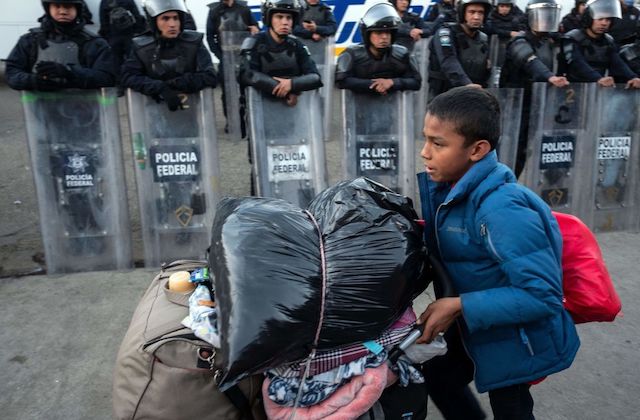 Young boy pushes his belongings in a grocery cart as he walks past a long line of Mexican officers
