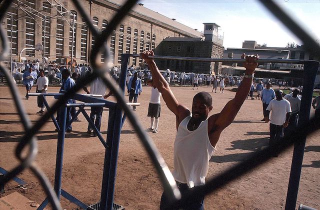 Black man in beige prison uniform does pull-ups outside in a prison yard.
