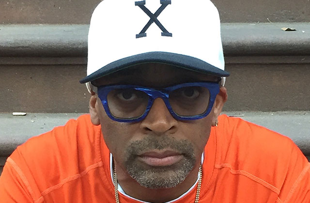 Spike Lee. Black man wearing white "X" cap with blue glasses and orange t-shirt.
