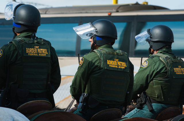 three US Border Patrol agents in green uniforms stand side by side with their backs to the camera.