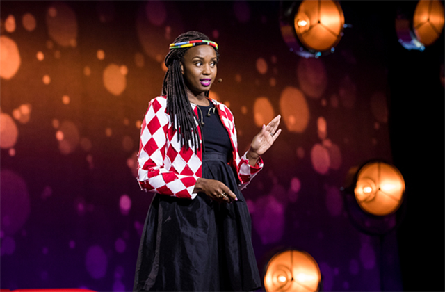 Wanuri Kahiu. Black woman with long braids wearing black dress and red-and-black checked jacket with beads around her head.