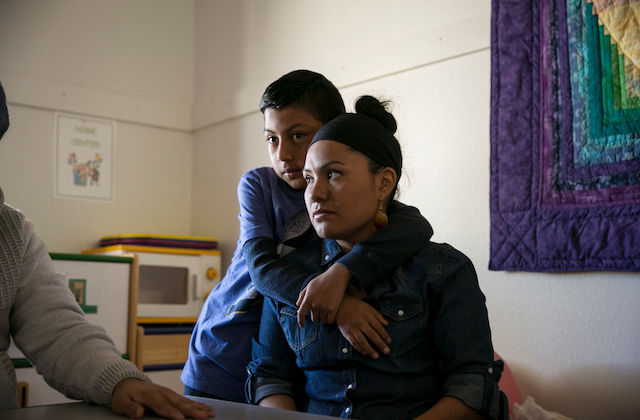 Latinx woman wearing denim shirt sits as her young son stands behind her with his arms wrapped around her shoulders.