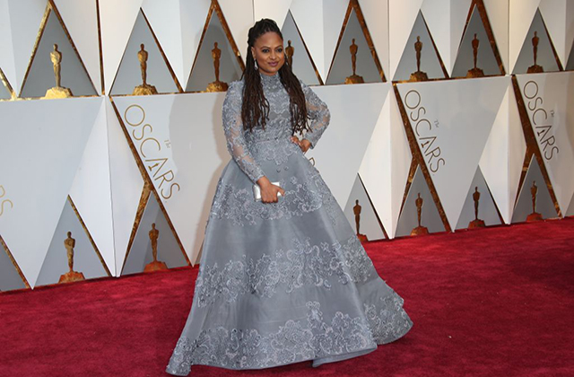 Ava DuVernay. Black woman with long brown locs wearing silver gown on red carpet standing in front of Oscar statues.