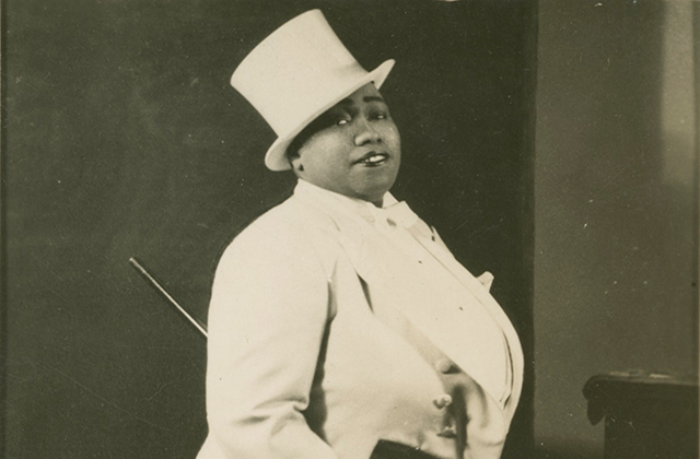 Gladys Bentley. Black woman wearing white top hat and white tuxedo with cane.