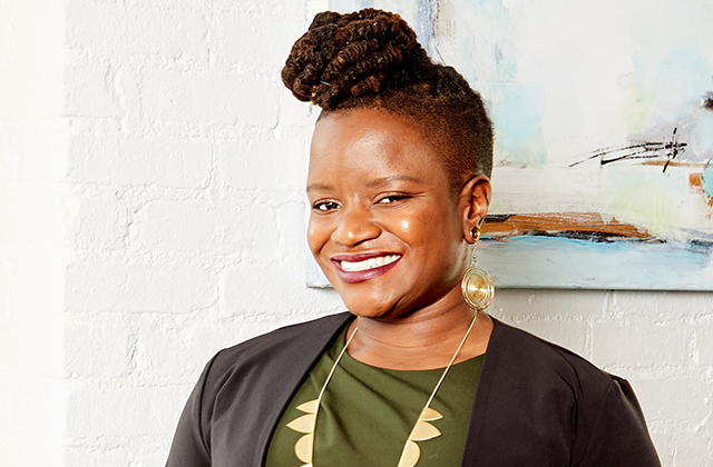 Chinyere Ezie. Smiling Black woman with locs in a high bun and shaved sides, wearing a green shirt, dark suit jacket and gold earrings and necklace.