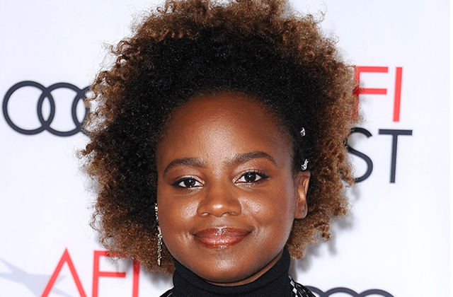 Dee Rees. Black woman with brown Afro, smiling, wearing a dark top in front of a step and repeat sign.