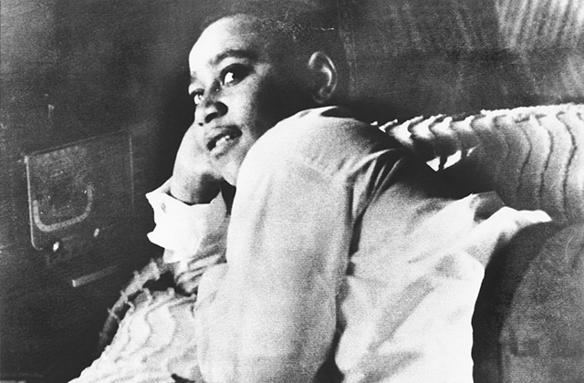 Emmett Till. Archival photo of young Black boy reclining on a bed with his head in right hand, wearing a white shirt and dark pants.