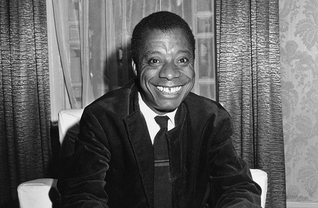 James Baldwin. Black man with short dark hair smiling broadly, wearing a white shirt and dark suit jacket, with dark tie.