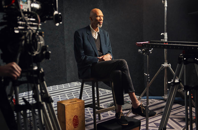 Kareem Abdul Jabbar. Slim older Black man with bald head and gray beard, wearing blue blazer, gray V-neck sweater and pants and a white button down top. He's sitting in chair with legs crossed surrounded by camera equipment.