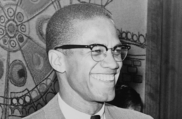 Malcolm X. Black man with short brown hair wearing glasses, white shirt, black tie, tan colored suit jacket, smiling.