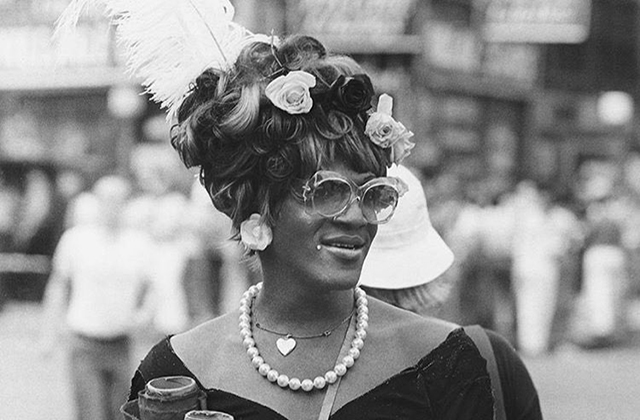 Marsha P. Johnson. Black woman with flowers and feather in hair, wearing glasses, flower earrings, pearl necklace and black sweetheart shaped top.