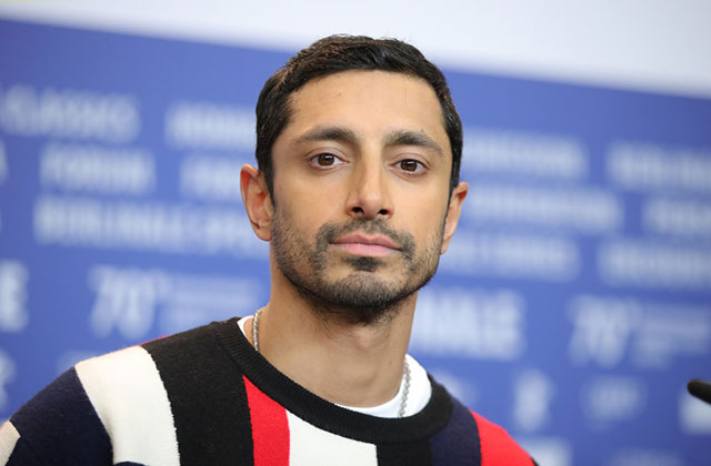 Riz Ahmed. South Asian man with short dark hair and short, groomed facial hair wearing red, white, blue colored sweater.