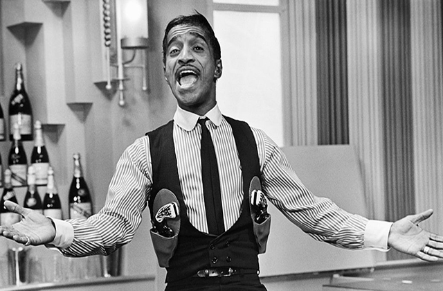 Sammy Davis Jr. Black man with short dark hair singing in a gun-slinger's outfit seated on a bar with his arms extended.