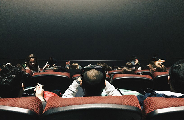 Movie theater. Dark room with brown chairs of people sitting rows facing  a dark theater screen.