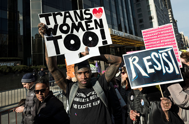 Protester. Black man at a march, wearing black T-shirt and gray backpack, holding a sign reading "Heart Trans Women Too!" next to sign that reads that "Resist."