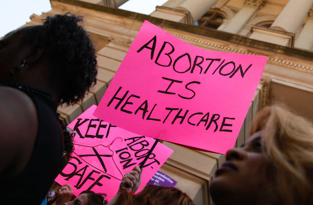 A pink protest sign that reads "Abortion is healthcare" is held at a rally in front of a government building