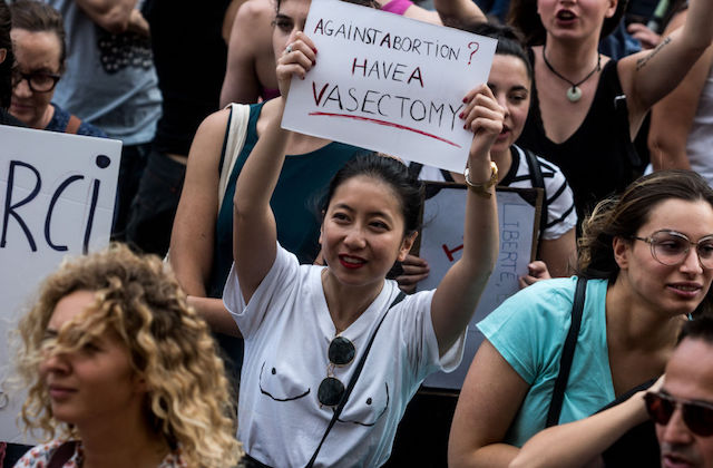 An Asian woman in a white t-shirt is at a protest and holds a sign that reads "Against abortion? Have a vasectomy."