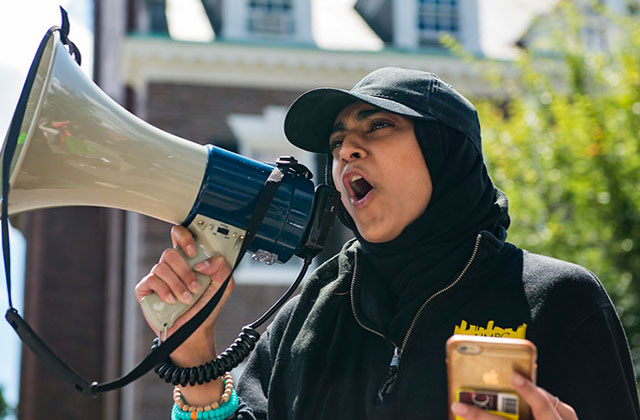 Anti-Hate Report. Muslim woman wearing a black cap over a black headscarf and black top holding a bullhorn.