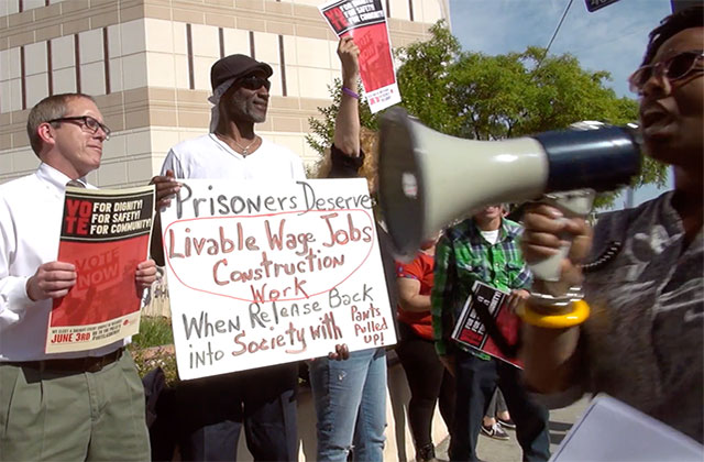 Scene from the documentary 'Bedlam' :  Protest scene where two men, one White, one Black, hold up signs and Black woman walks by speaking on a microphone.