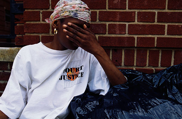 COVID-19-Domestic Violence. Young Black woman looking distressed in front of a brick building covers her face, wearing a colorful headscarf and white T-shirt, standing behind black garbage bags.