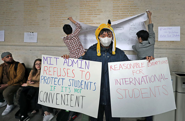 Coronavirus protest. Asian-American student wearing yellow hat, white face mask, blue coat holding up to two protest signs.