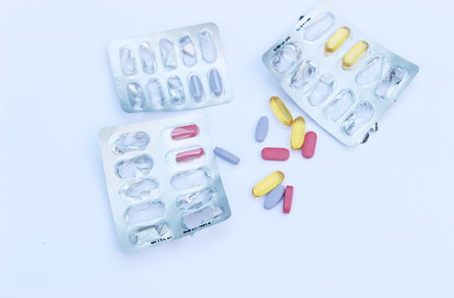 Pills. Pink, blue, and yellow pills next to empty pill packets on a desk.