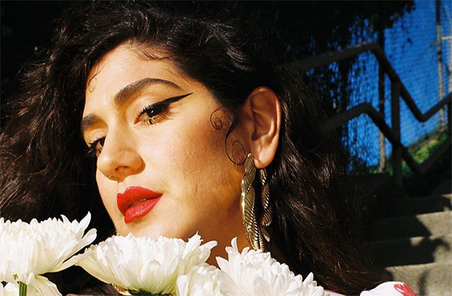 La Dona. Latinx woman with long dark hair wearing red lipstick, large hoop gold earrings, in front a row of white flowers.