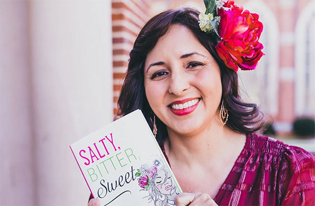 Mayra Cueva. Latinx person with a red flower in their hair, wearing a red top and holding up a children's book.