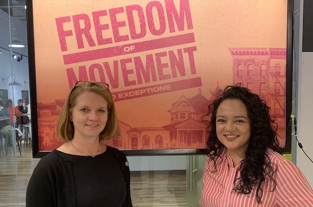 Two women stand in front of an orange sign that reads "Freedom of Movement"