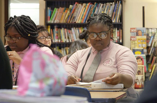 PUSHOUT. Two teenage Black girls in a classroom with book shelves behind them as they sit at a desk wearing pink button up shirts, gray ties, long braids and glasses.