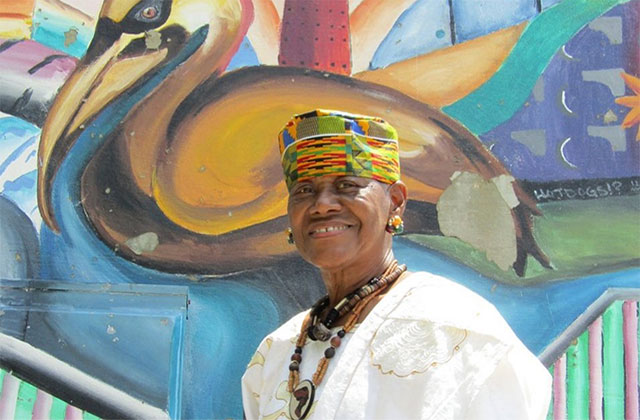 Sadie Roberts Joseph. Older Black woman wearing colorful hat with white top and brown beaded necklace, standing in front of colorful wall mural.
