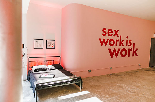Sex Workers Popup. Pink wall reads "sex work is work" next to a bed.