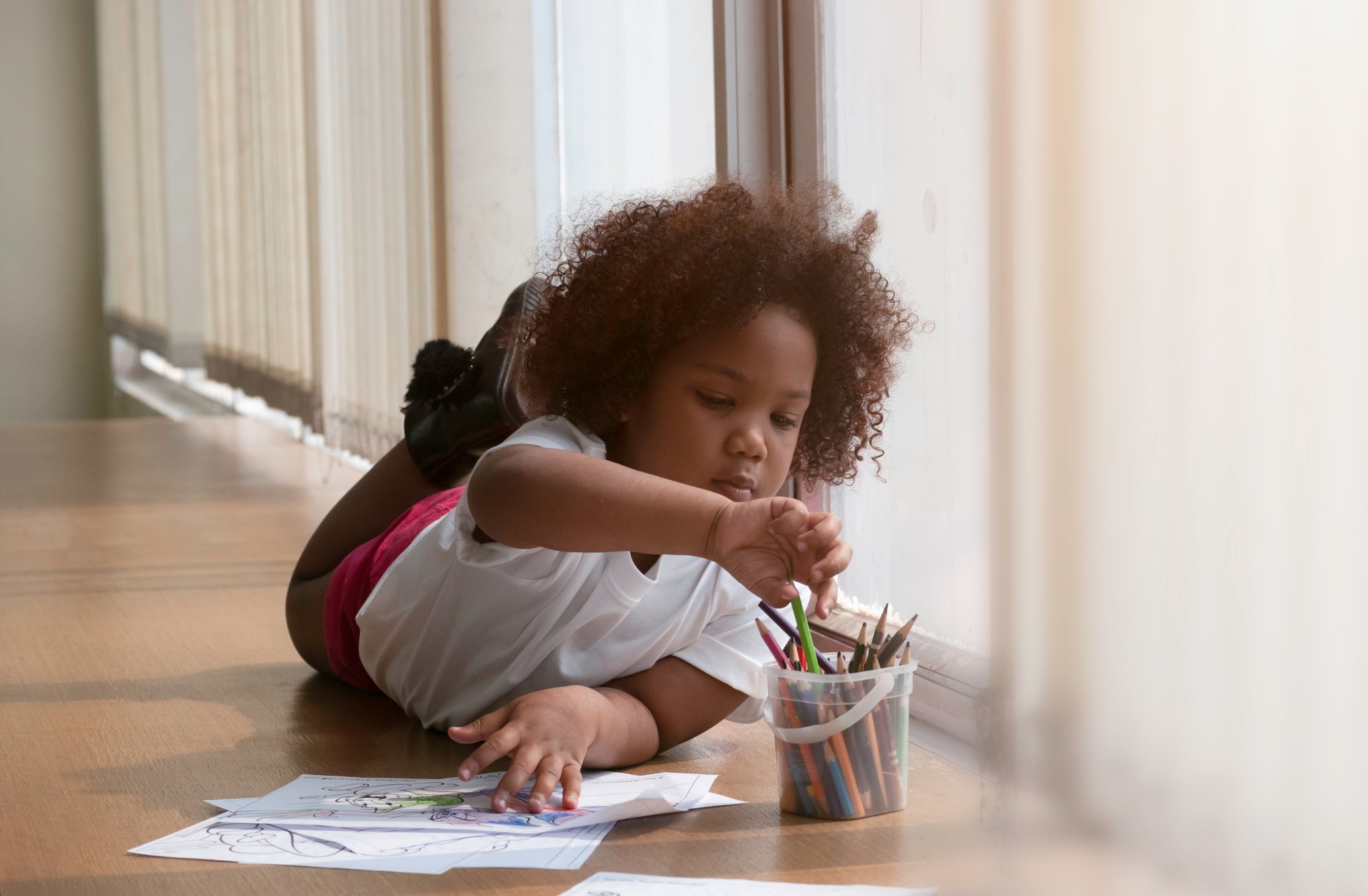 School age African American girl laying on the floor of sunny room with paints