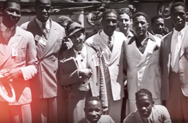 1936 Olympics. Archival image of about nine Black men and women huddled together for a photo with everyone wearing a suit.