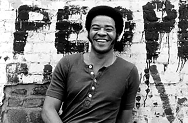 Bill Withers. Black and white photo with Black man in a short Afro and dark short sleeve t-shirt standing in front of brick wall with "People" spray painted on it.