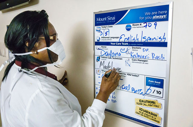 Black nurse. Black woman with dark hair wearing a white mask and lab coat writing at a schedule board.