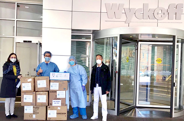 Four people standing in front of Wycoff Hospital and around many brown boxes.