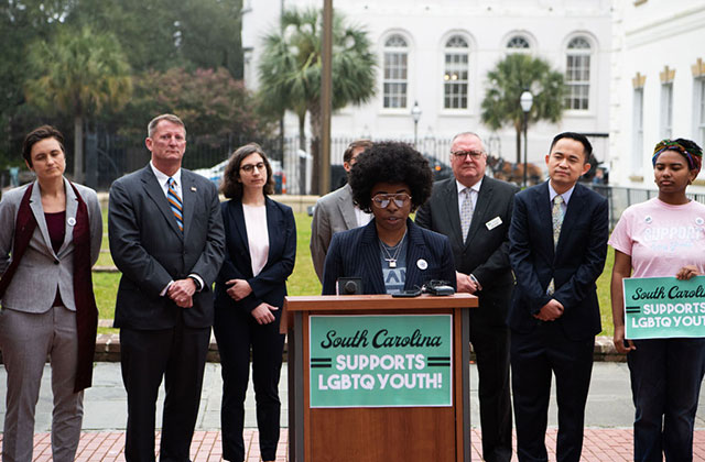 Campaign for Southern Equality. Black woman with Afro wearing a dark suit and classes stands outside in front of wooden podium, in front a row of people, also in suits.
