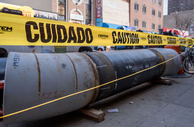 Yellow caution tape surrounds rows of pipeline left on the side of an urban street.