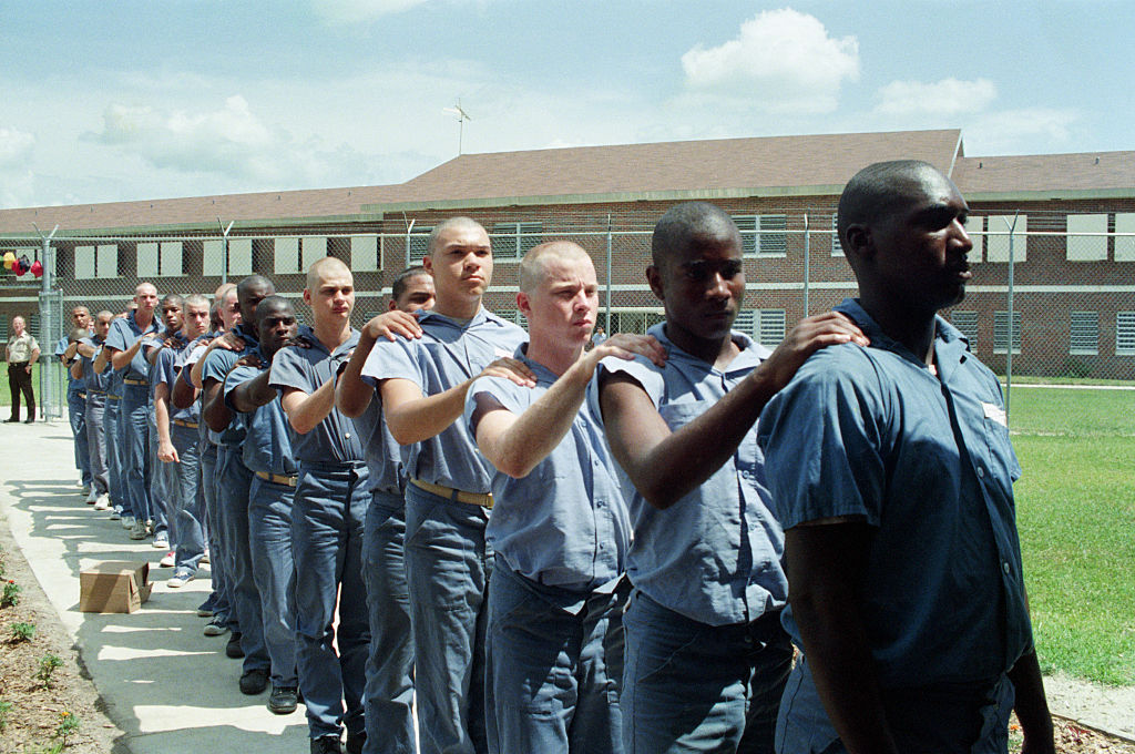 Single file line of bald headed men, black and white in blue prison uniforms lined up with one hand on the person's shoulder in front of them.