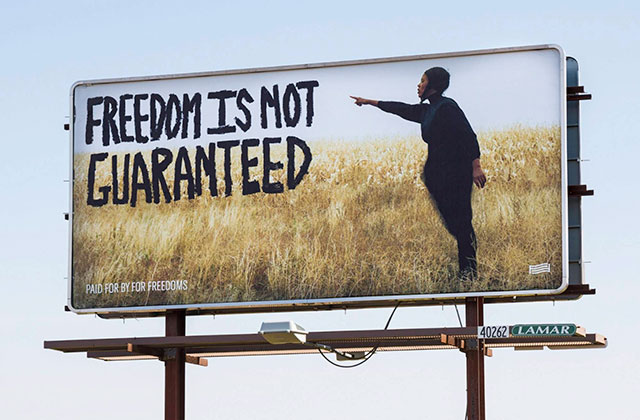 Billboard. Photo of a young Black man standing in a field of tall grass pointing to words in big black letters that read: "Freedom Is Not Guaranteed."